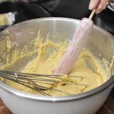 Sumergir cada salchicha en la masa, ayudarse con una cuchara si es necesario. Colocar cada una en un barquito de papel aluminio untado con mantequilla para que no se peguen y poner los barquitos sobre una charola para horno.