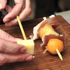 Armar las brochetas colocando primero un cubo de queso, después una uva, un cuadro de jamón serrano y la bola de melón. Repetir hasta tener la brocheta completa.